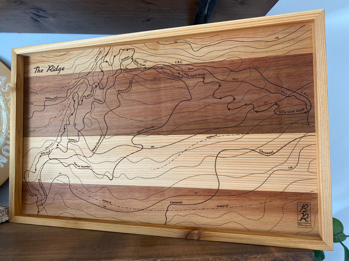 A wooden trail map of The Ridge on Forbidden Plateau with an engraved design showcasing a topographic outline, displayed in a frame.