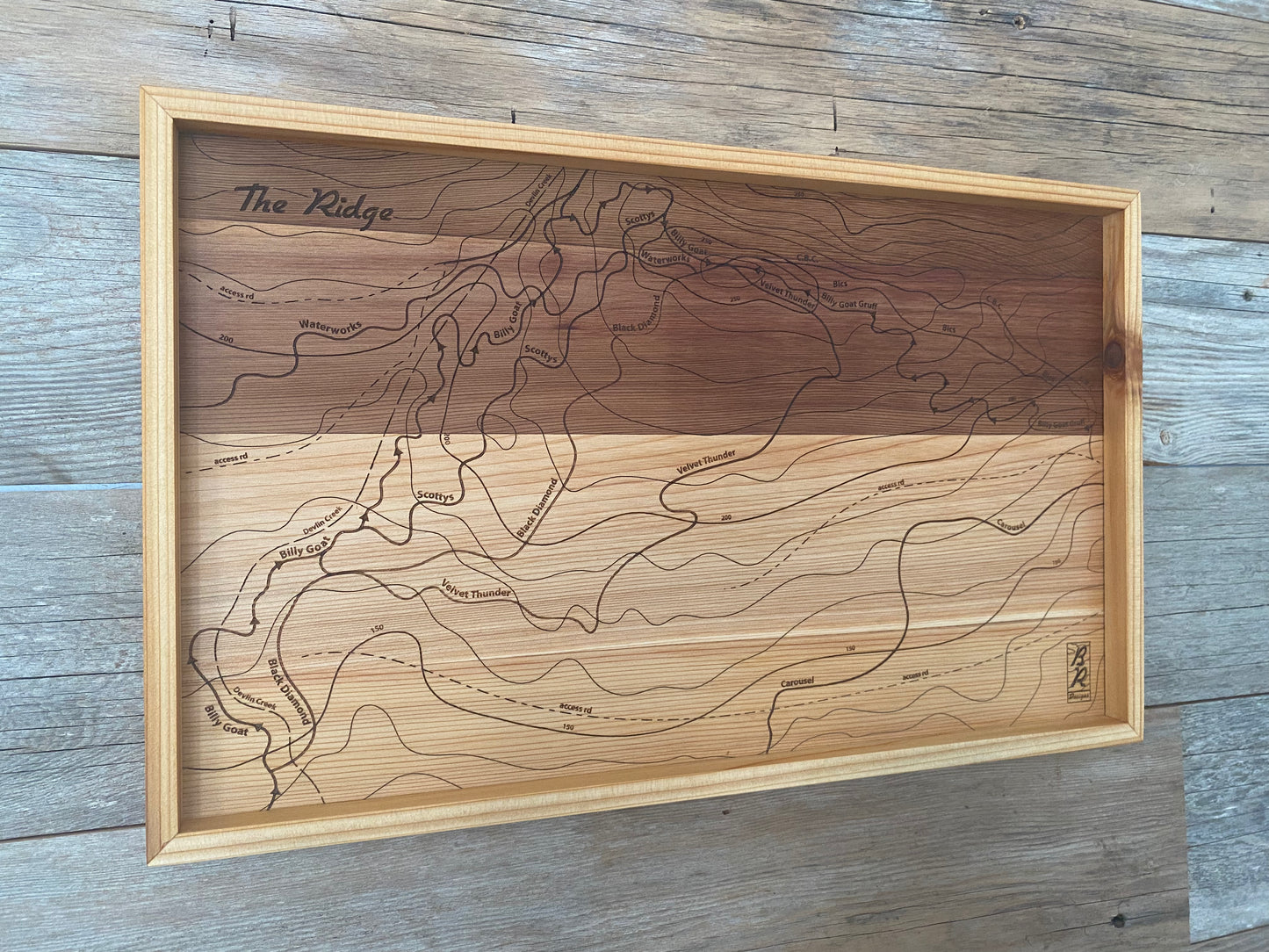 A wooden trail map of The Ridge on Forbidden Plateau with an engraved design showcasing a topographic outline, displayed in a frame on a wood wall