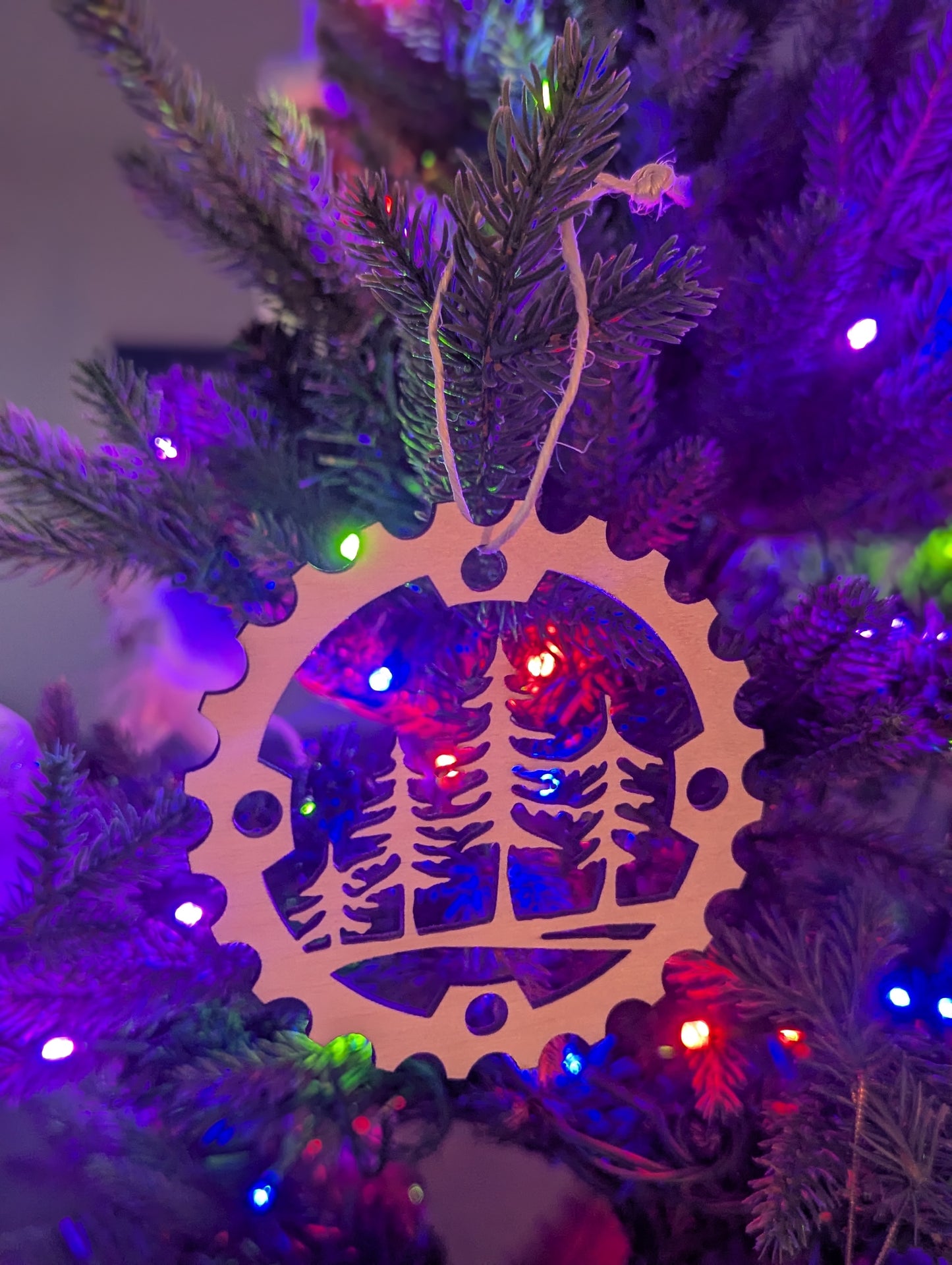 A Christmas tree ornament designed like a mountain bike chain ring with a tree scene in the center, hanging from a tree with colorful lights and a natural hanger.