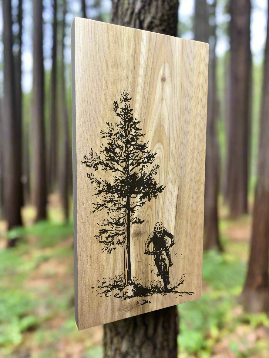 A wood wall tile with a mountain biker and pine tree design, hanging in a forest.